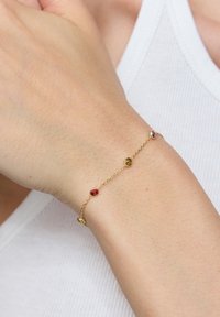 Złota bransoletka z trzema kolorowymi kamieniami: czerwonym, żółtym i fioletowym, na delikatnym łańcuszku, spoczywająca na nadgarstku o jasnym odcieniu skóry.