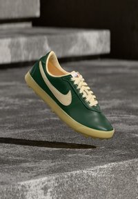 Πράσινο και κρεμ αθλητικό πα πούτσι Nike με κορδόνια, που αιωρείται πάνω από υφή πέτρινων σκαλοπατιών, ρίχνοντας σκιά στο έδαφος από κάτω.