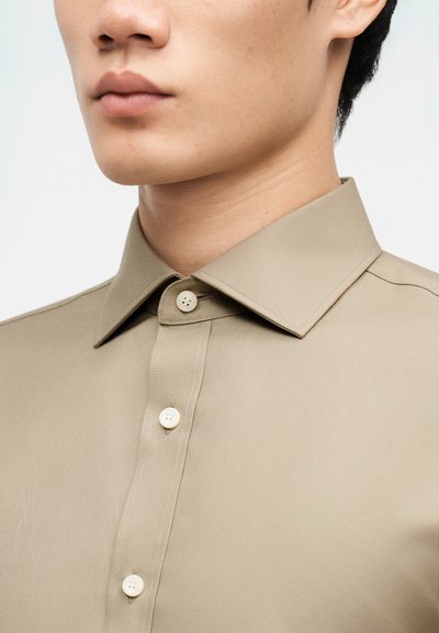 Κοντινό πλάνο ενός ατόμου που φοράει μπεζ πουκάμισο με κουμπιά και μυτερό γιακά, δείχνοντας το κάτω μέρος του προσώπου, το λαιμό και το πάνω μέρος του στήθους.