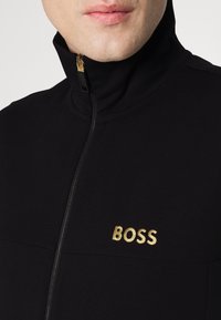 Černá bunda na zip s vysokým límcem, zdobená zlatě vyšívaným logem "BOSS" na hrudi a hladkým materiálem. Viditelný zlatý zip.