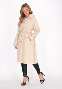 Beige trenčkot s páskem v pase, zapínán�í na knoflíky a velkým límcem. Obsahuje boční kapsy a dlouhé rukávy, stylizovaný s černými legínami a podpatky.