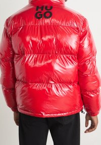 Rote Steppjacke mit glänzendem Finish, gesteppter Optik, dicker Polsterung und dem Markenzeichen "HUGO" in Schwarz auf dem oberen Rücken.