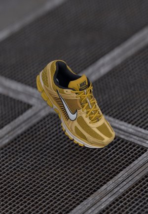 Gelber Nike Laufschuh mit schwarz-weißem Logo, dargestellt über Metallgitterboden.