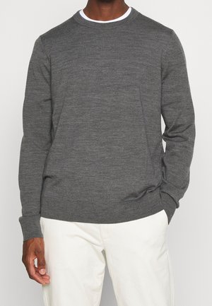 Mand iført en mørkegrå crewneck-trøje over en hvid skjorte, kombineret med hvide bukser, stående med den ene hånd i lommen.