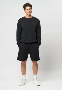 Set din sudor negru și pantaloni scurți, cu accente roșii ale logo-ului. Materialul pare moale și are o croială lejeră. Pantofii sport și șosetele albe completează ținuta.
