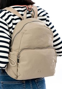 Beige rugzak met een gladde textuur, twee met rits afsluitbare compartimenten en een logodetail. Wordt gedragen door een persoon met een gestreept shirt en spijkerbroek.