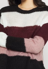 Pletený svetr s horizontálními pruhy v černé, bílé, burgundské a růžové barvě. Volná pletená textura s kulatým výstřihem a dlouhými rukávy.