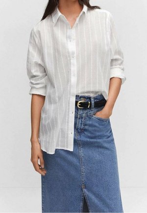 Vrouw draagt een wit textuurrijk overhemd met opgerolde mouwen, ingestopt in een blauwe denim rok met een zwarte riem met gouden gesp.