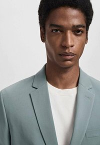 Blazer grigio chiaro realizzato in tessuto liscio, con revers a punta e tasca sul petto singola, indossato sopra un maglione girocollo bianco.