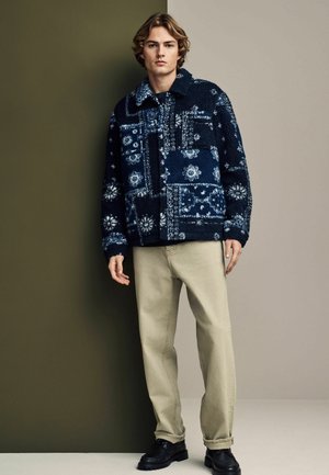 Tummansininen kuviollinen fleece-takki, jossa on kauluksellinen kaula-aukko ja suuri etutasku, yhdistettynä vaaleanbeigeihin housuihin ja mustiin kenkiin.