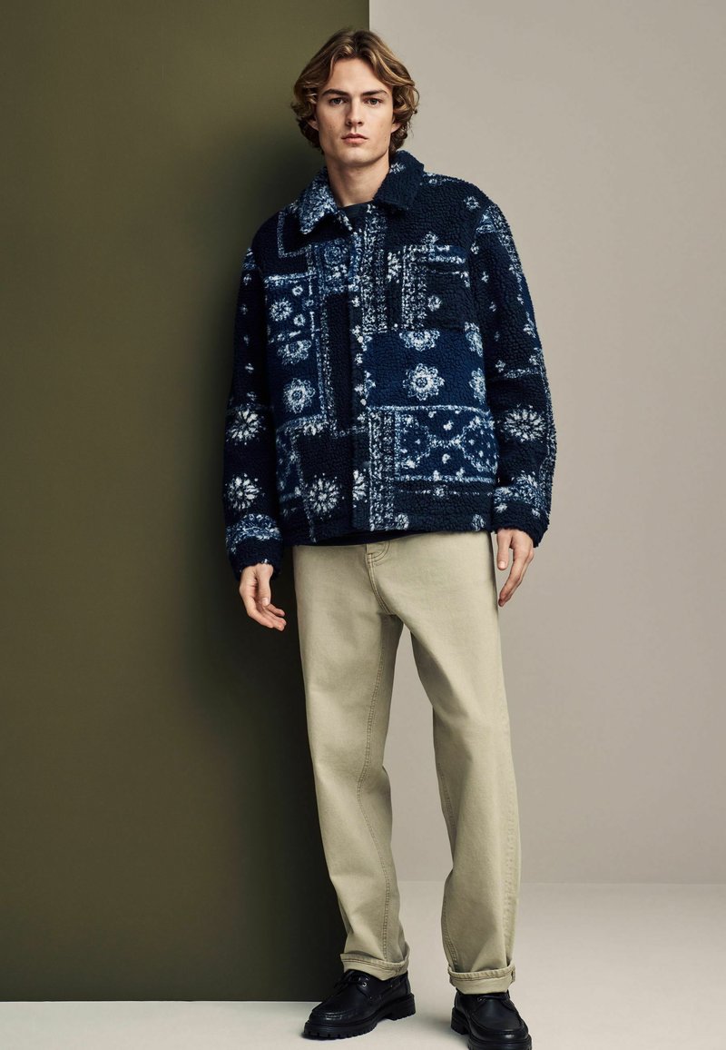 Marineblauwe fleece jas met een kraag, grote voorkantzak, gecombineerd met lichtbeige broek en zwarte schoenen.