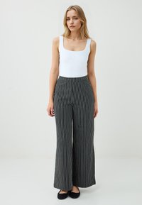 Een model staat zelfverzekerd, gekleed in een witte tanktop en zwart-wit gestreepte, wijde broek, tegen een effen lichte achtergrond.