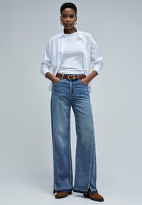 Wide-leg light blue jeans paired with a white t-shirt and a white button-up shirt. Brown leather belt and boots. Smooth denim texture.