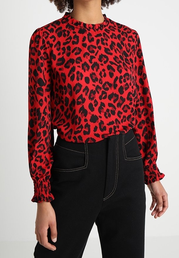 Femme portant un chemisier rouge à manches longues avec des taches léopard noires et un pantalon noir taille haute avec des détails de couture blancs.