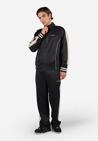 Sort tracksuit med cremeaccenter, med lynlåslukning og elastiske manchetter; matchende bukser med sidelister. Sorte træningssko fuldender looket.