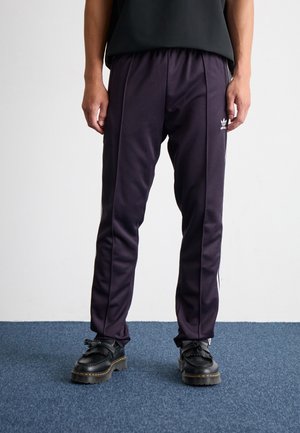 Osoba stojící na modrém koberci, oblečená v tmavě fialových teplácích Adidas s bílými pruhy a černých masivních botách.