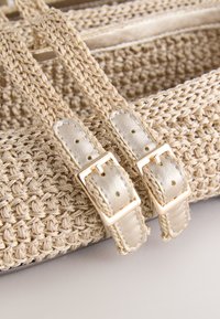 Gros plan sur des anses de sac tissées beige avec boucles en métal doré et accents en cuir sur un sac texturé assorti.