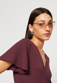 Sunglasses with a transparent orange lens, sleek metal frame, and elongated shape. The subject has straight dark hair and wears a burgundy top.