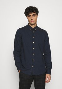 Marineblaues Hemd aus strukturiertem Stoff mit spitzem Kragen, Holzknöpfen und langen Ärmeln. Kombiniert mit gemusterten Hosen.