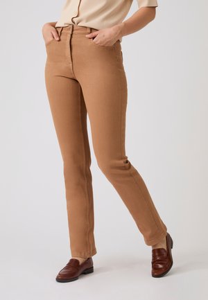 Persona che indossa una camicia beige abbottonata, pantaloni slim fit color cammello e mocassini in pelle marrone, in piedi con le mani in tasca.