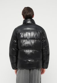 Jachetă puffer neagră cu un finisaj lucios, având un guler înalt și secțiuni cu matlasare orizontală. Textură moale, căptușită, cu o croială lejeră.