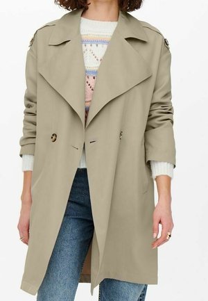 Trench coat beige con design doppiopetto, ampio colletto e dettagli con bottoni; abbinato a un maglione in maglia e jeans.