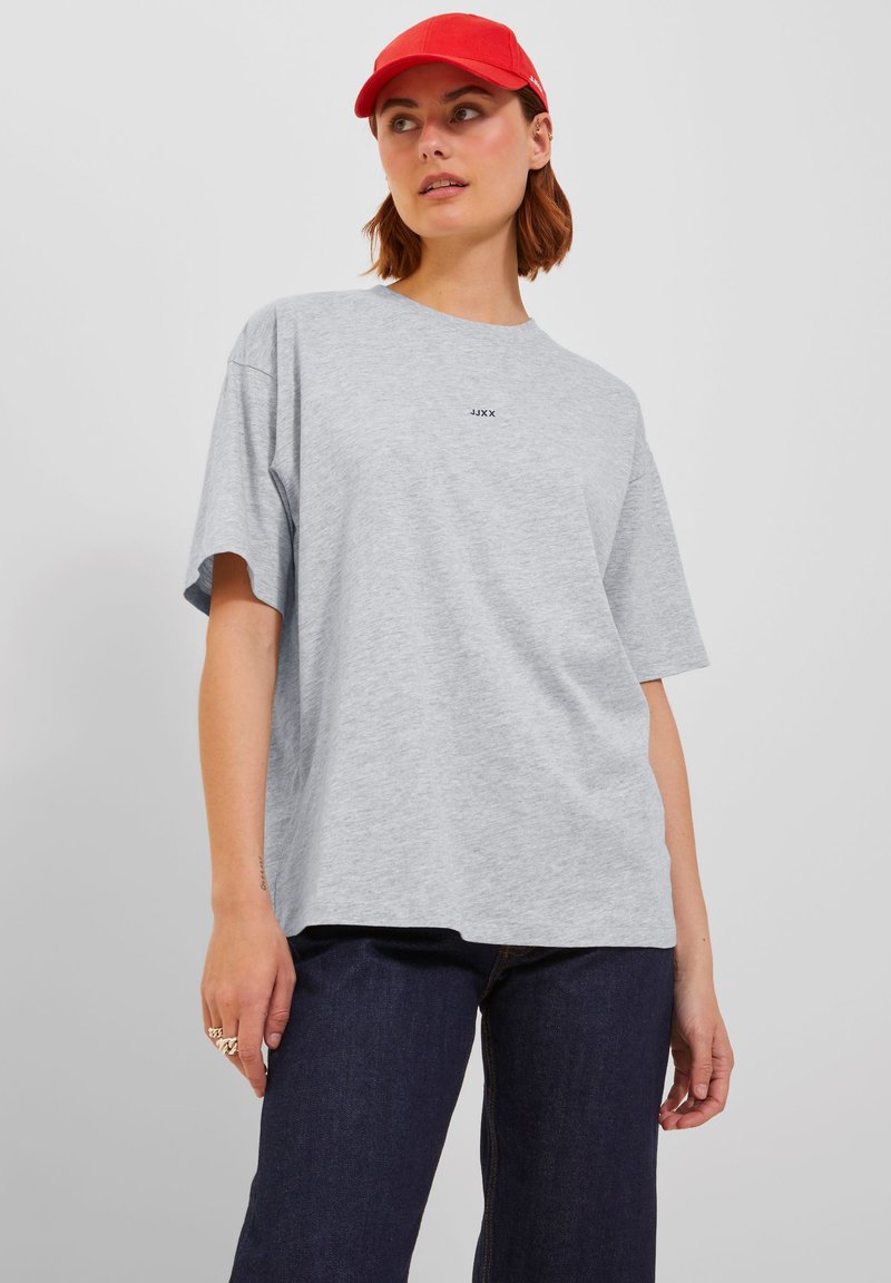 Grå bomull T-skjorte med korte ermer og løs passform, med en svart logo på brystet. Kombinert med mørk denimbukser og en rød cap.