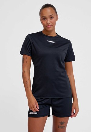 Kobieta w czarnych sportowych ubraniach HUMMEL, w tym koszulce z krótkim rękawem i szortach, stoi z widocznymi tatuażami na ramionach i nodze.