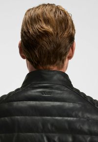 Zwarte leren jas met een horizontaal gestikt patroon, voorzien van een kraag en een subtiele "BOSS"-logo op de achterkant. Glad materiaal, aangesloten ontwerp.
