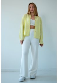Żółty sweter z dzianiny, prążkowany biały crop top oraz białe rozszerzane spodnie. Sweter ma guziki i luźny krój, o gładkiej fakturze. Białe trampki.