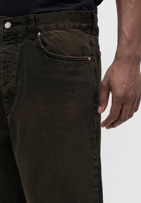 Κοντινή λήψη τσέπης και μέσης ανοιχτό καφέ τζιν με ορατές ραφές και χαλαρό δεξί χέρι ενός ατόμου δίπλα της.
