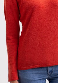 Röd stickad tröja med texturerad yta, långa ärmar och rundad nederkant, kombinerad med blå denimjeans.