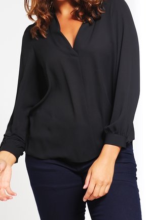 Woman wearing a black long-sleeve blouse with a V-neck and dark pants, with curly hair partially visible.