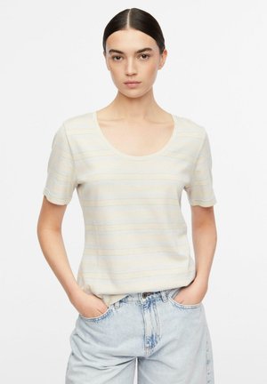 Frau mit dunklen Haaren in einem cremefarben gestreiften Kurzarmshirt und hellblauen Jeans, steht mit den Händen in den Taschen vor weißem Hintergrund.