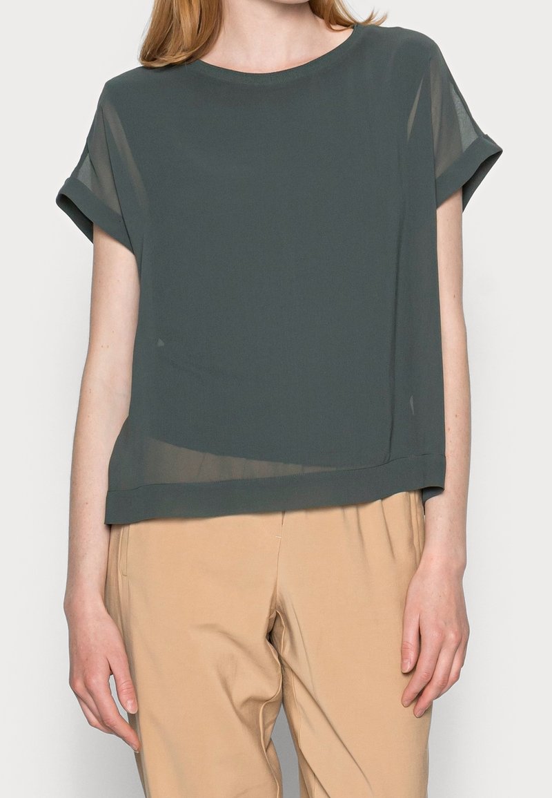 Femme portant un chemisier court en voile vert foncé à manches courtes retroussées et un pantalon beige taille haute sur un fond uni.
