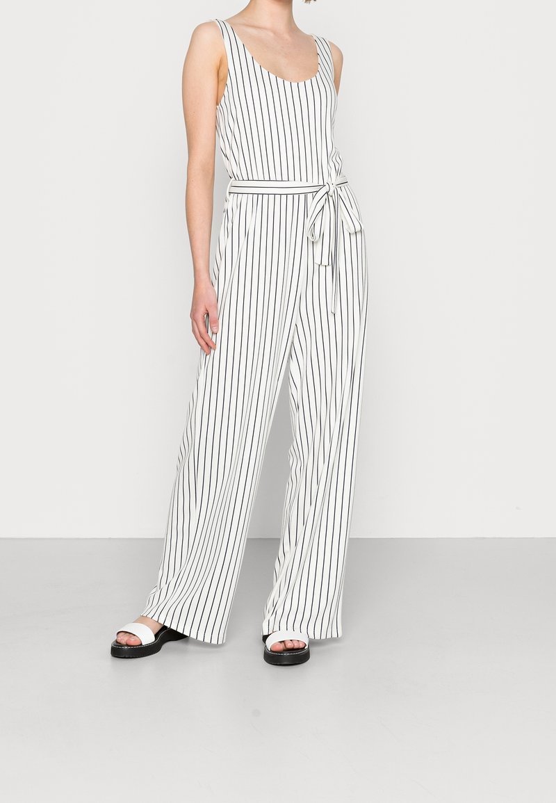 Femme portant une combinaison sans manches blanche à rayures verticales noires, taille nouée et pantalons larges, associée à des sandales blanches.