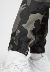 Camo-Jogginghosen in Grautönen und Schwarz mit elastischen Bündchen an den Knöcheln. Kombiniert mit weißen Sneakers und knöchelhohen Socken.
