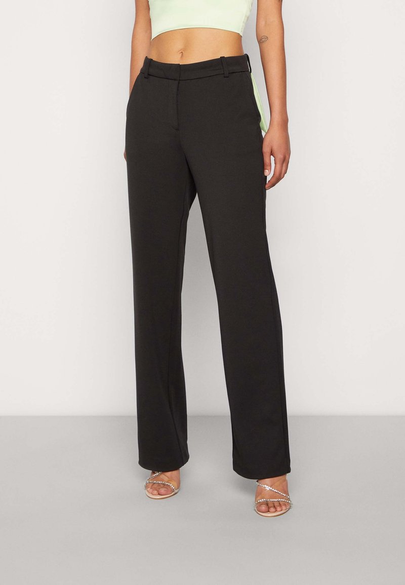 Femme portant un pantalon noir taille haute à jambes larges, associé à des sandales à talons argentées à brides et un top court vert clair.