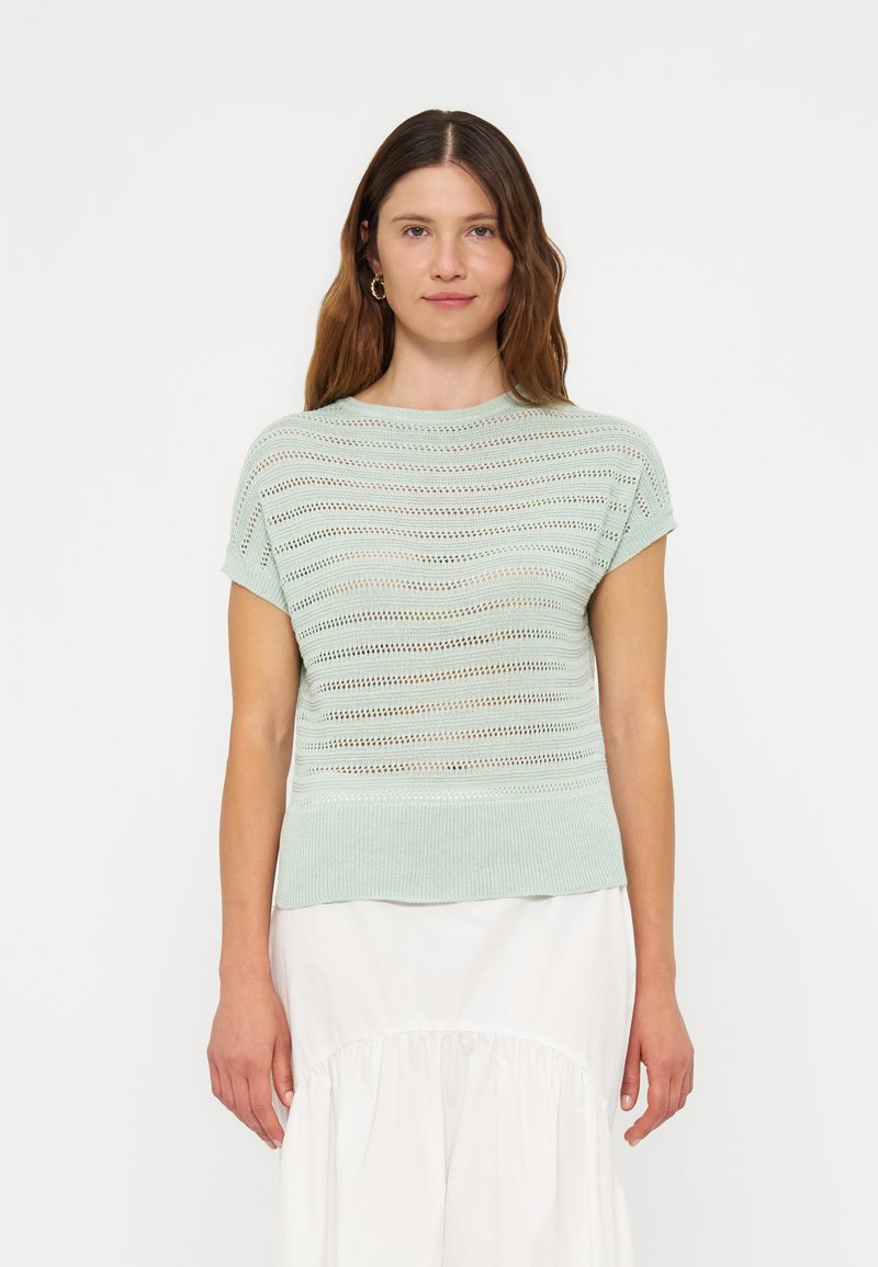 Femme aux longs cheveux bruns portant un haut en maille vert clair à manches courtes et une jupe blanche, debout devant un fond blanc uni.