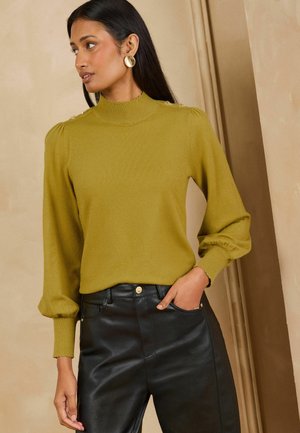 Femme portant un haut jaune moutarde à manches longues avec des détails de boutons sur les épaules et un pantalon en cuir noir, debout à l'intérieur avec une main dans la poche.