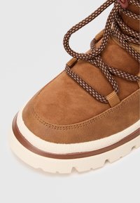 Bottine en daim marron avec une finition texturée, dotée d'une semelle en caoutchouc marron et crème, et de lacets marron à motifs passés à travers des œillets en métal.