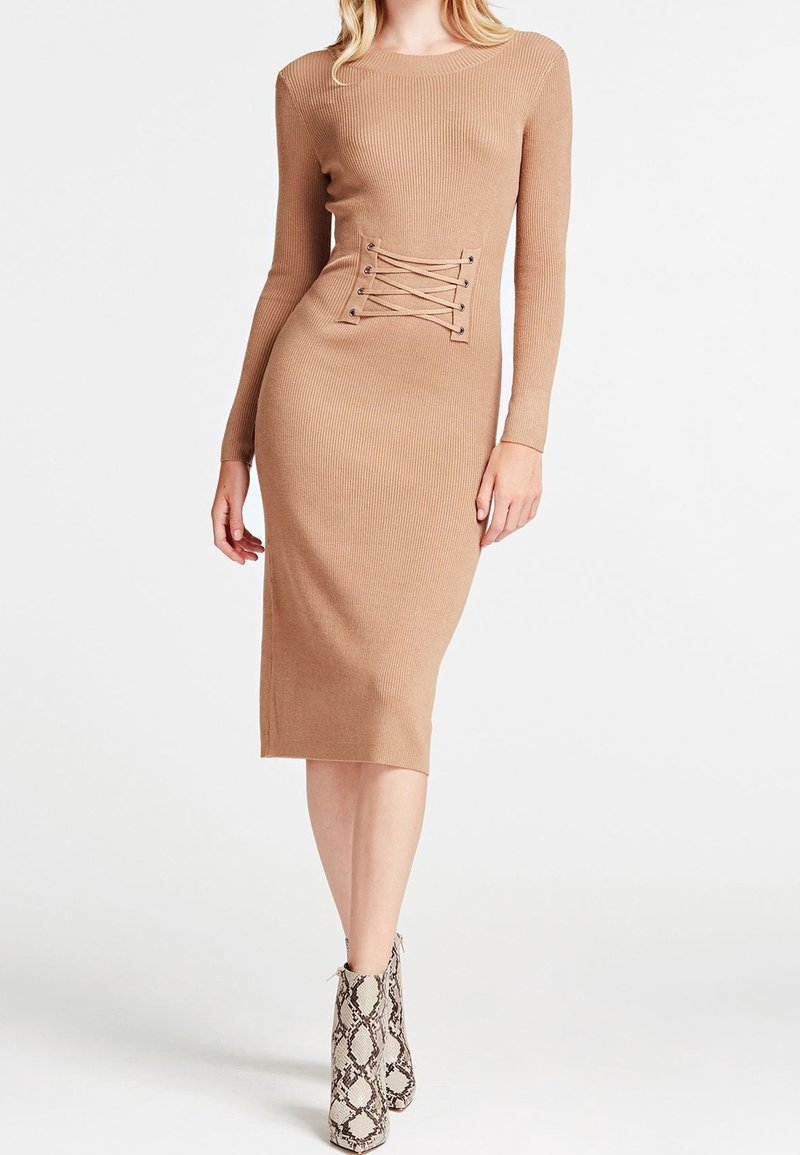 Femme portant une robe midi camel ajustée à manches longues, avec un détail lacé à la taille et des bottines en cuir serpent.