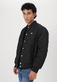 Svart quilted bomberjakke med trykknapper, ribbede mansjetter og hem, samt en liten logoaccent på brystet. Glatt stoffstruktur.