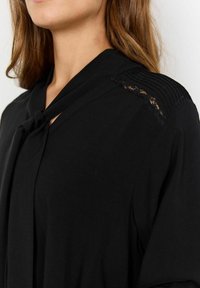 Blusa negra con un lazo anudado en el frontal, detalles de pliegue en el hombro y acentos de encaje en el escote. Tela suave y ligera.