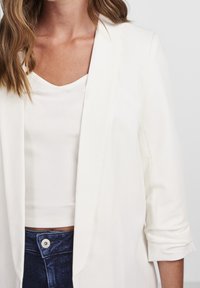 Blazer blanc avec une texture lisse et un design structuré, porté sur un haut blanc côtelé, assorti à un pantalon bleu marine taille haute.