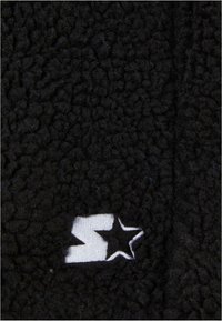 Sort fleece stof med en tekstureret, fluffy overflade. Har et hvidt logo patch med en stjerne og et stiliseret bogstav 'S'.