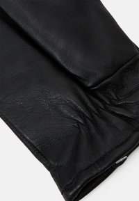 Gant en cuir noir avec une surface douce et légèrement texturée. Le poignet est froncé pour un ajustement confortable, mettant en avant un design épuré avec un minimum de matériel.
