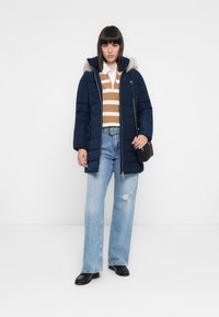 Cappotto puffer blu navy con cappuccio in pelliccia staccabile, maglione a righe marroni e bianche, jeans a gamba larga chiari e strappati, e borsa a spalla nera.
