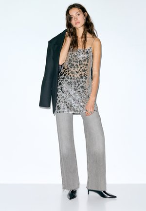 Zilveren glitterjurk met spaghettibandjes en een zwarte blazer. Grijze wijde broekspijpen en zwarte schoenen met puntige neus maken de outfit compleet. Eenvoudige achtergrond.