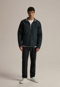 Navy blue pikowana kurtka z zamek błyskawicznym i białym paskowanym kołnierzykiem; noszona na białej koszulce, w zestawie z ciemnymi spodniami i białymi trampkami.